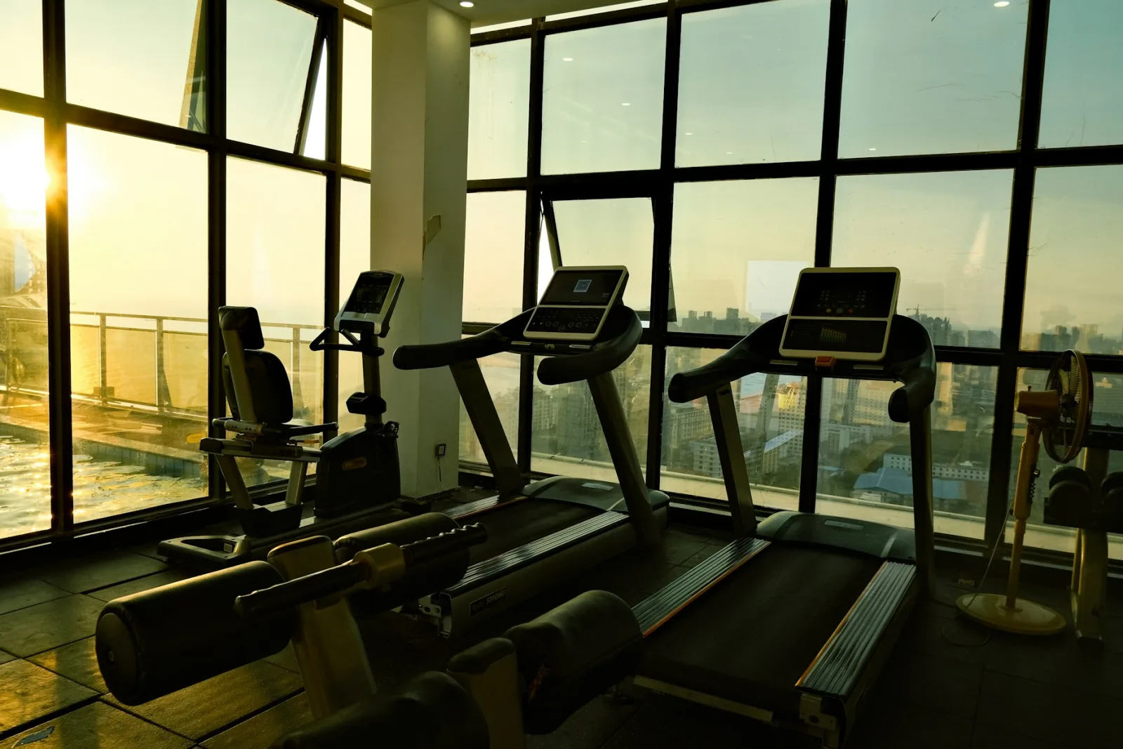 Phòng gym có máy chạy bộ xếp cạnh cửa kính lớn nhìn ra thành phố